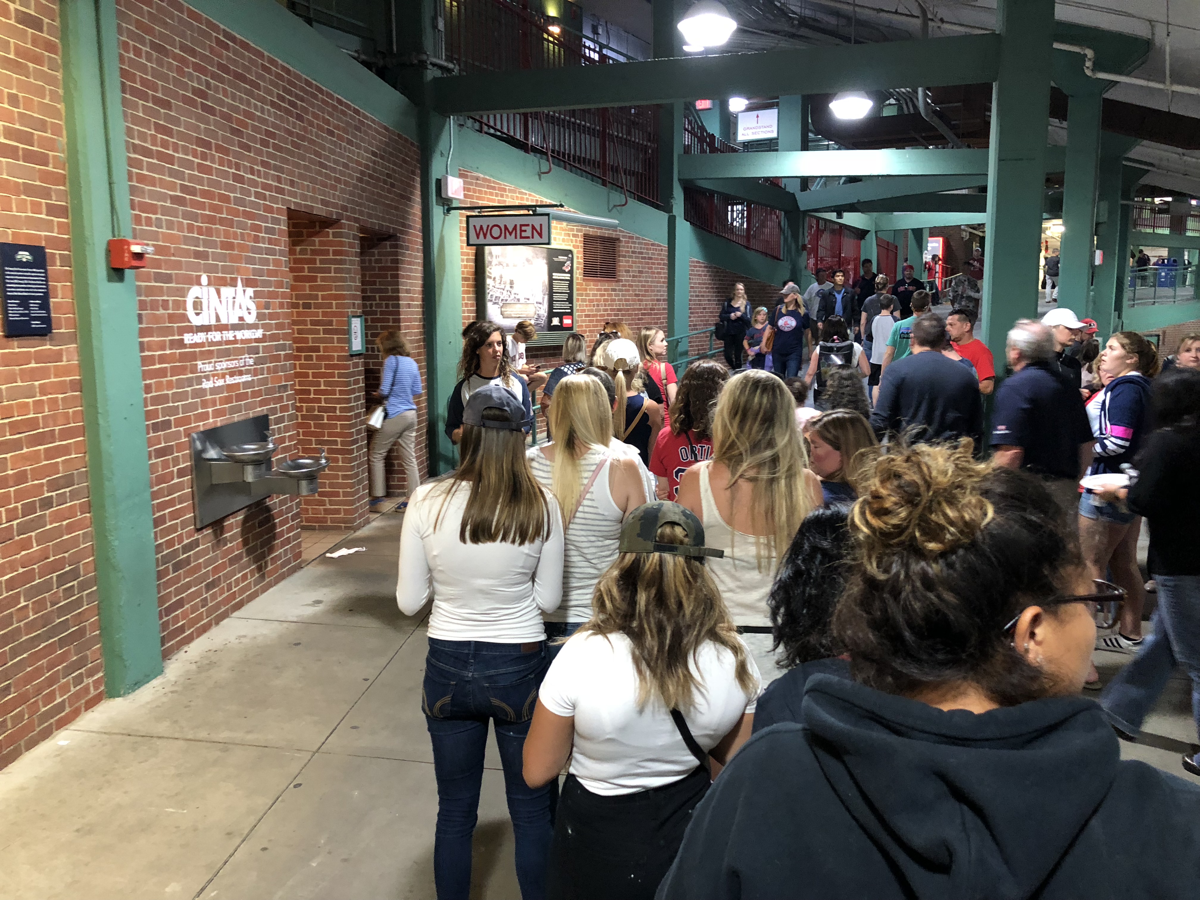 a line of about 10 women dressed in casual and Boston Red Sox attire, waiting in line outside the bathroom