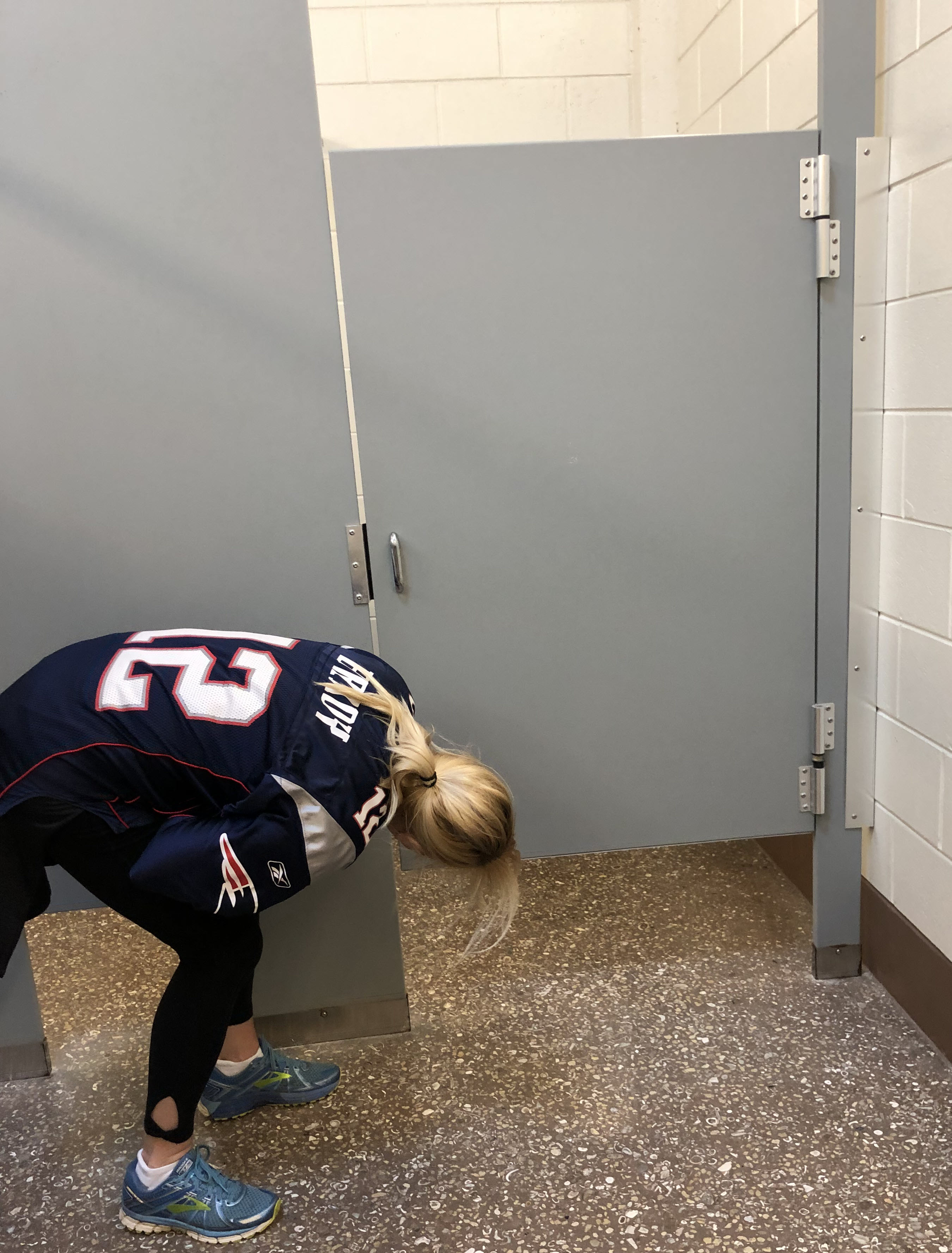 a blonder woman in a New Englad Patriots jersey leans down outside a stall to look for feet