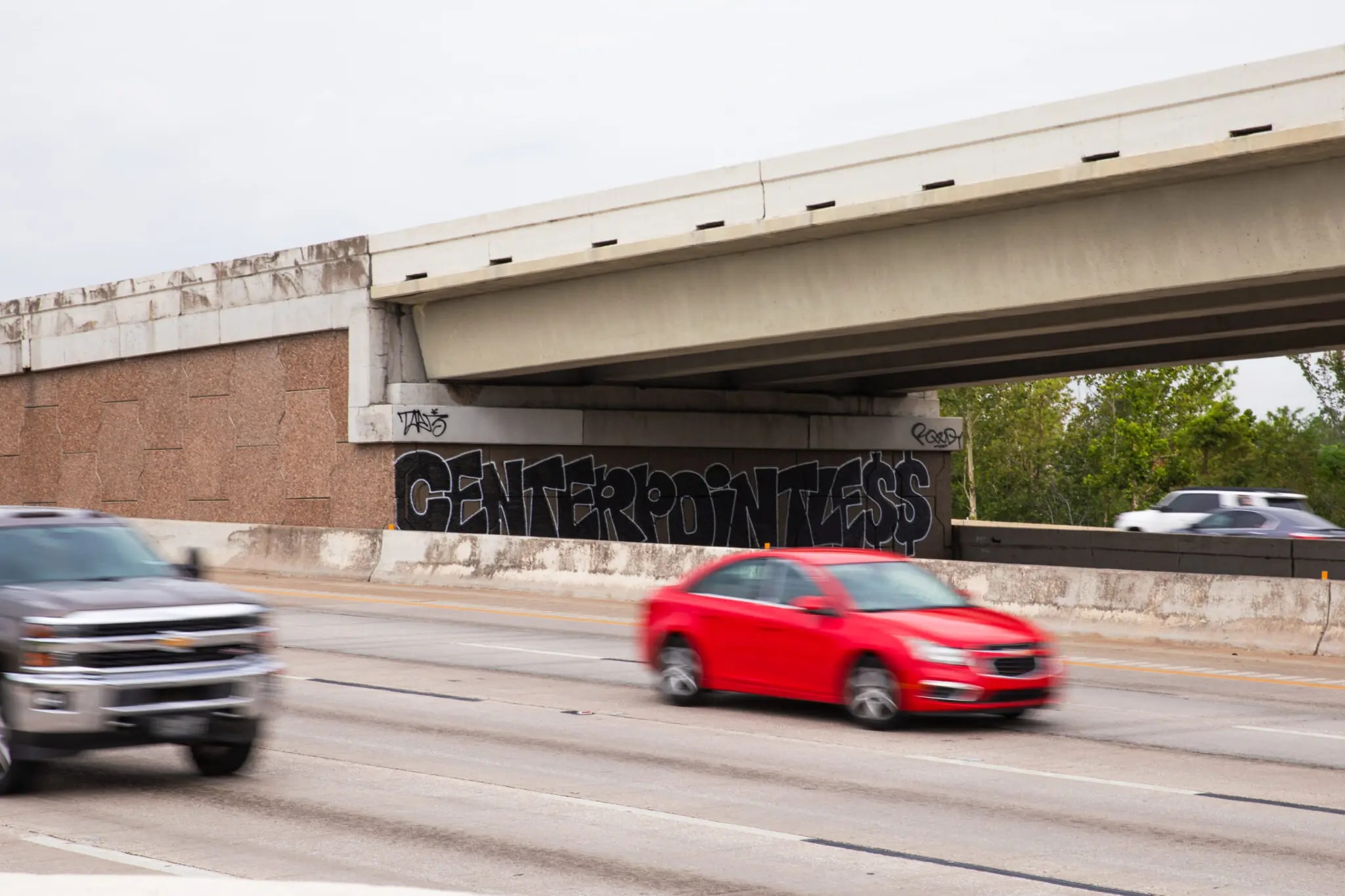 a section of a highway with an overpass. On the concrete support of the overpass, there is large graffiti that reads “CENTERPOINTLESS” in black, bold letters. The graffiti appears to be a critical commentary, likely directed at CenterPoint Energy. Several cars are driving on the highway, including a red car and a gray truck in the foreground, with other vehicles visible in the background. The overall scene suggests movement and urban environment. The weather appears to be overcast, adding a muted tone to the setting.