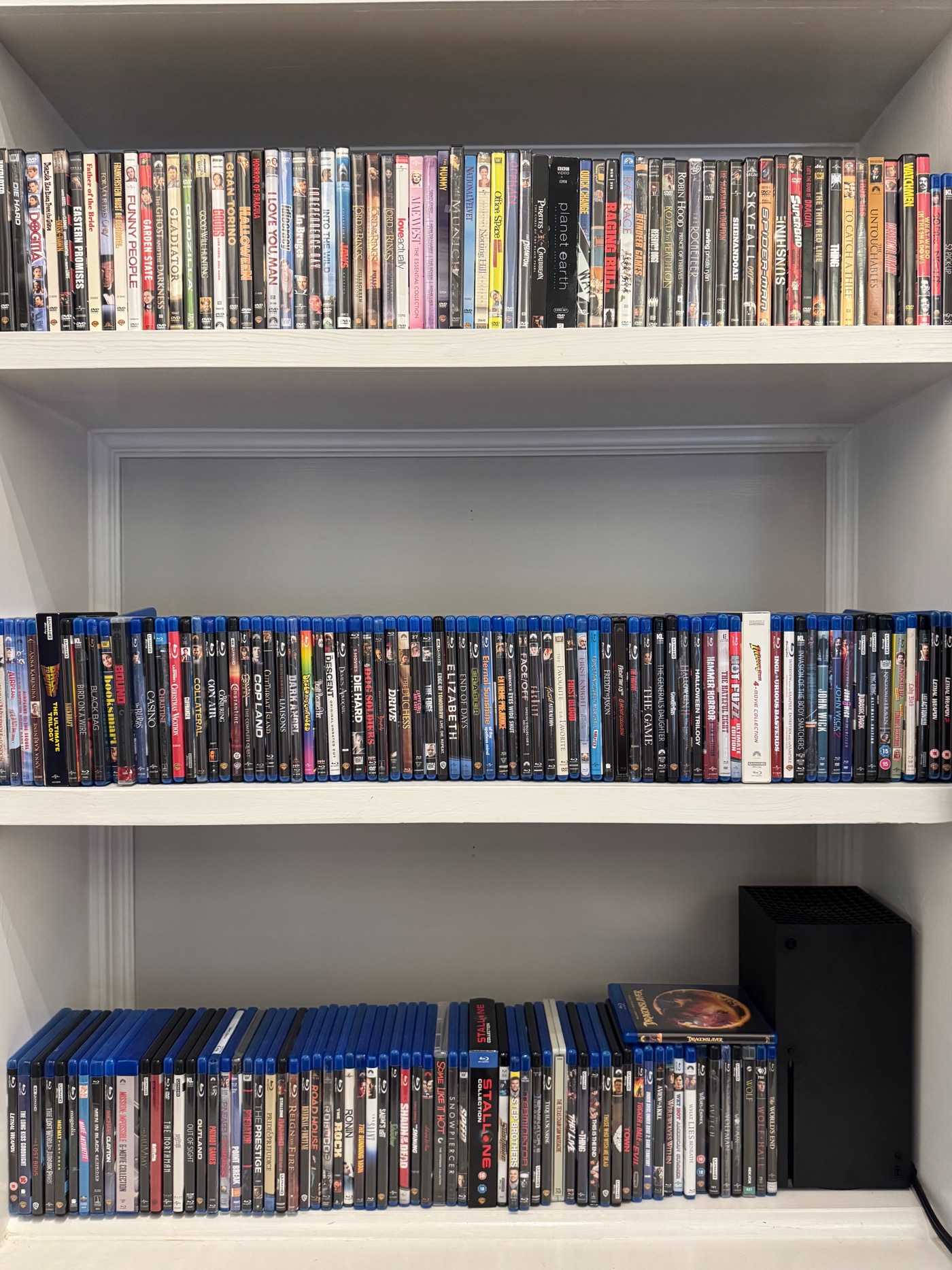 Tres bastidores con DVD y Blu-ray, y una consola Xbox en la rejilla inferior. Las grandes colecciones físicas representan el regreso del autor al disco en la transmisión.